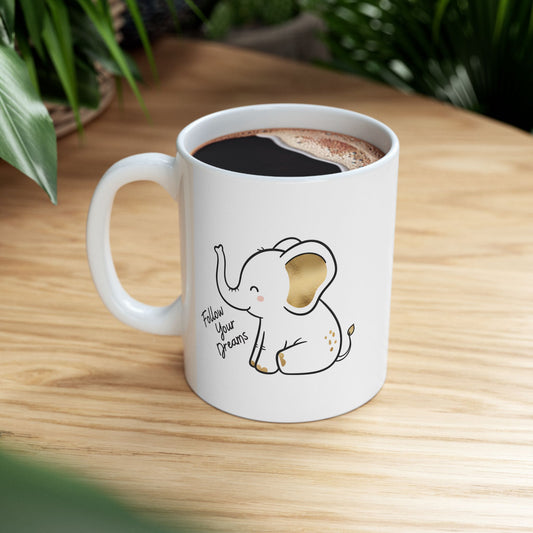 White mug with an elephant illustration and 'Follow Your Dreams' text, filled with a dark liquid on a wooden surface.