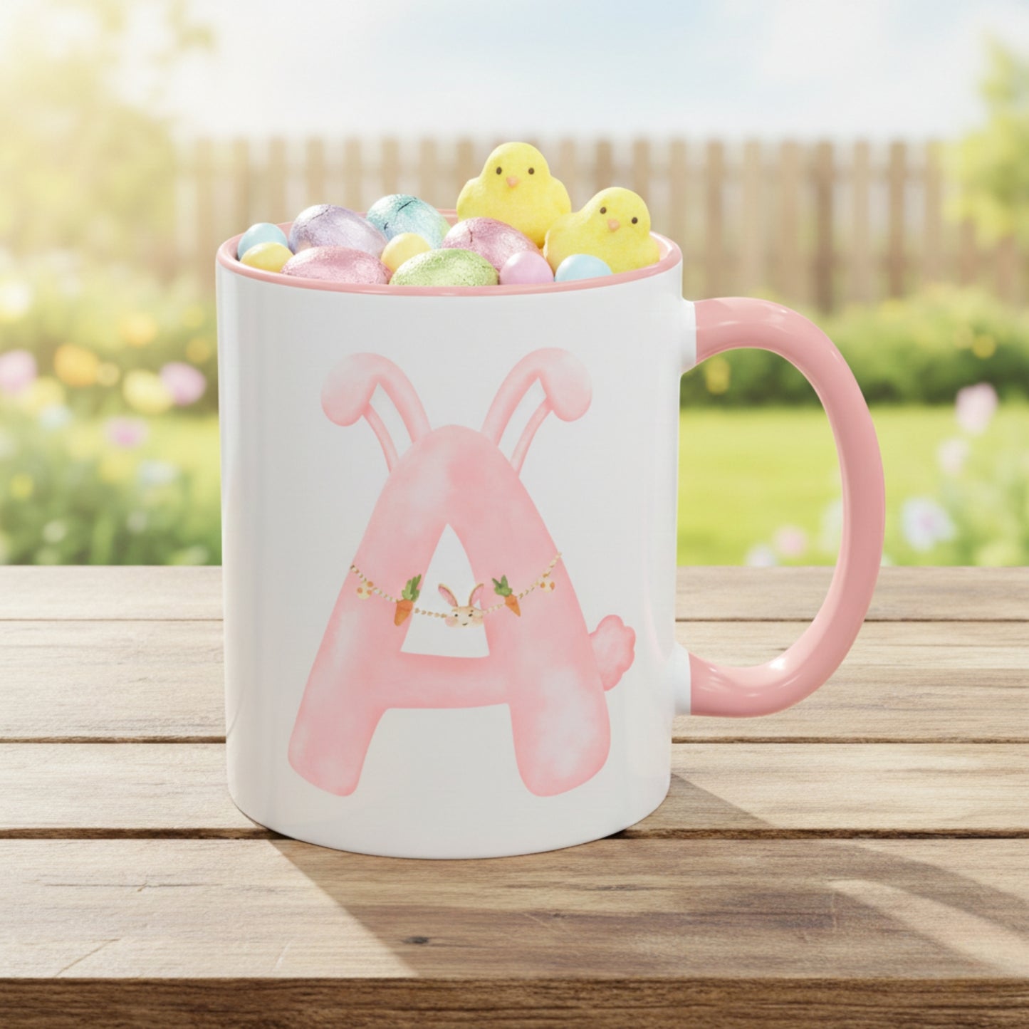 White mug with pink letter 'A' design filled with colorful chocolate Easter eggs and marshmallow chicks on a wooden table outdoors.